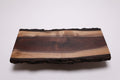 A rectangular live-edge black walnut wooden board with a polished dark brown center and natural rough bark edges, photographed on a clean white background.