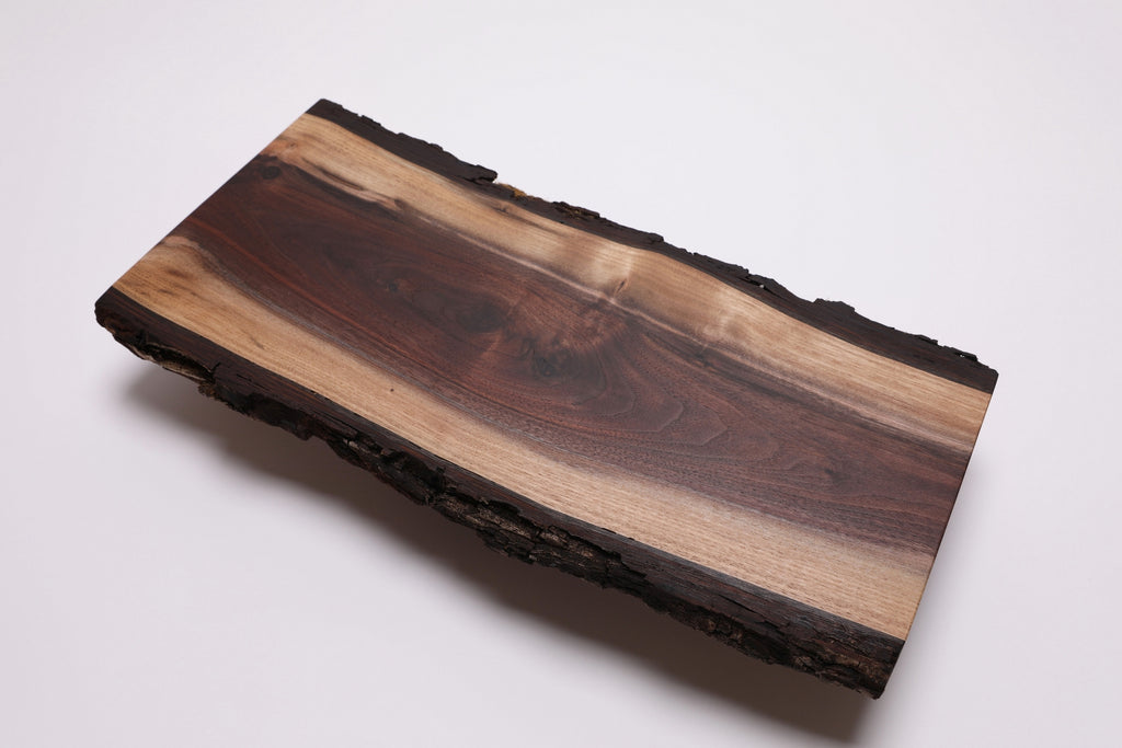 A rectangular live-edge black walnut wooden cheese/charcuterie board with a natural bark border, featuring a rich wood grain pattern in dark brown and light tan tones, photographed on a clean white background.