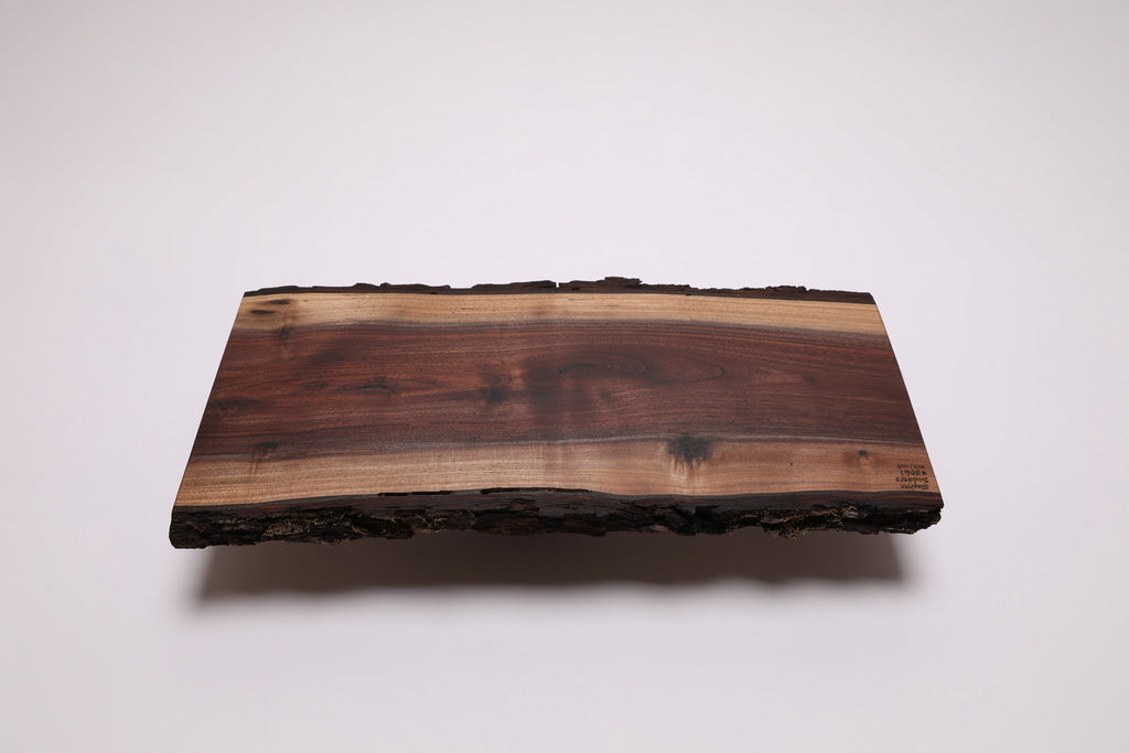 A rectangular live-edge wooden board with a natural bark border, featuring a rich gradient of wood tones from dark brown to light tan, shown on a clean white background.