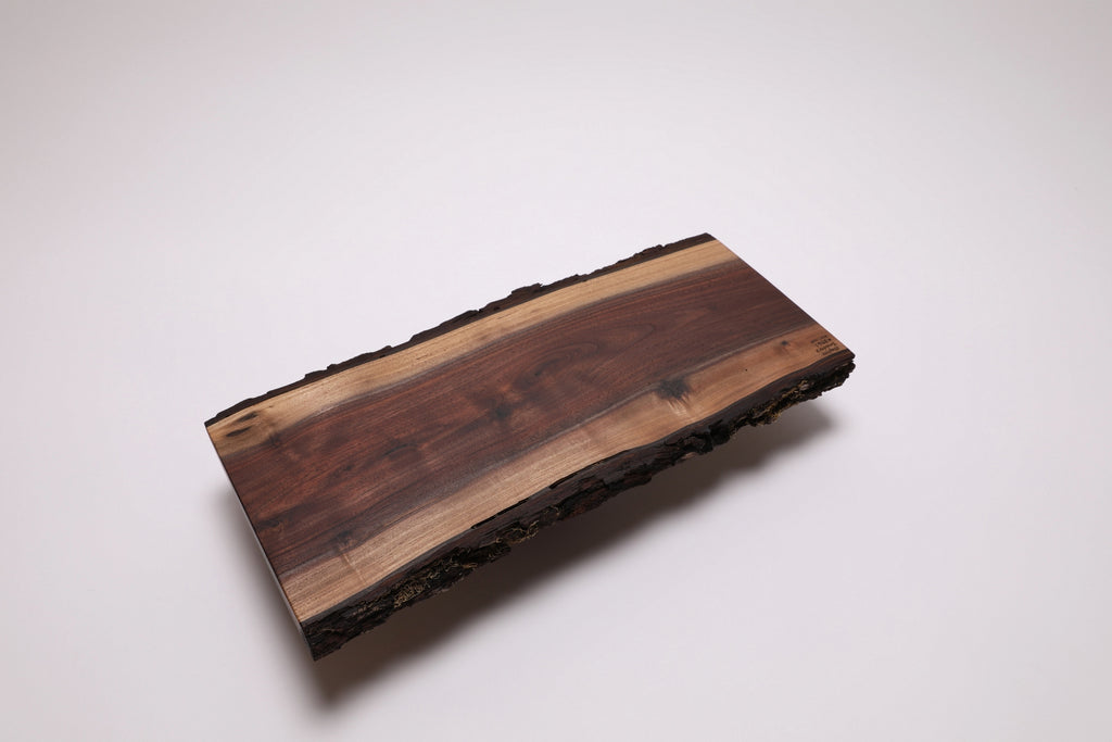 Bottom view of a rectangular live-edge black walnut wooden serving board with a natural bark border, featuring a dark brown center and lighter tan wood grain, photographed on a clean white background.