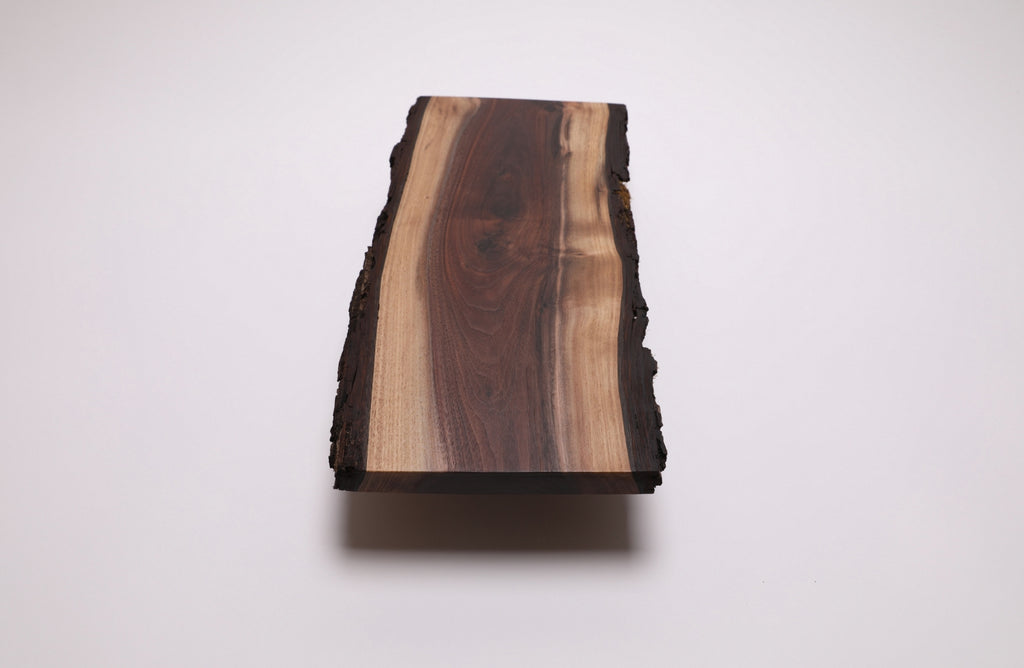 A rectangular live-edge walnut wooden board with a natural bark edge, featuring a prominent dark brown wood grain center and lighter tan wood grain on the sides, shown on a plain white background.