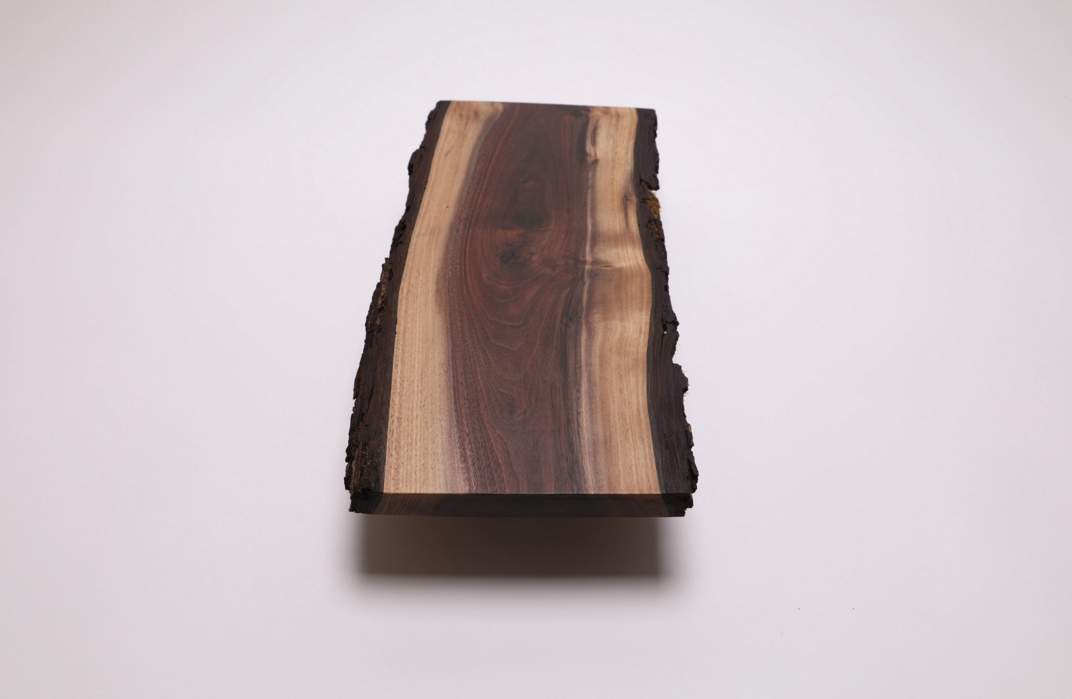 A rectangular live-edge walnut wooden board with a natural bark edge, featuring a prominent dark brown wood grain center and lighter tan wood grain on the sides, shown on a plain white background.