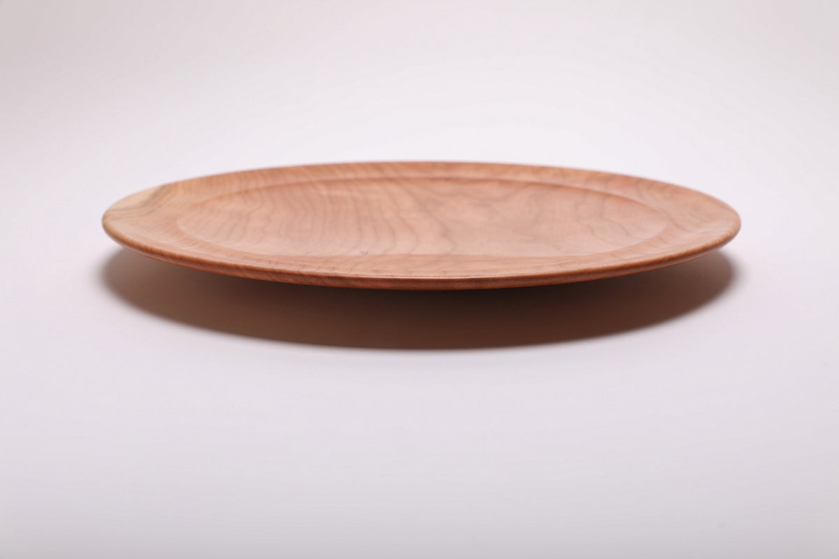 Side profile of hand-turned curly wild black cherry wooden platter showing thin curved taper, raised foot, and warm pink-toned exterior, 1 1/2 inch height