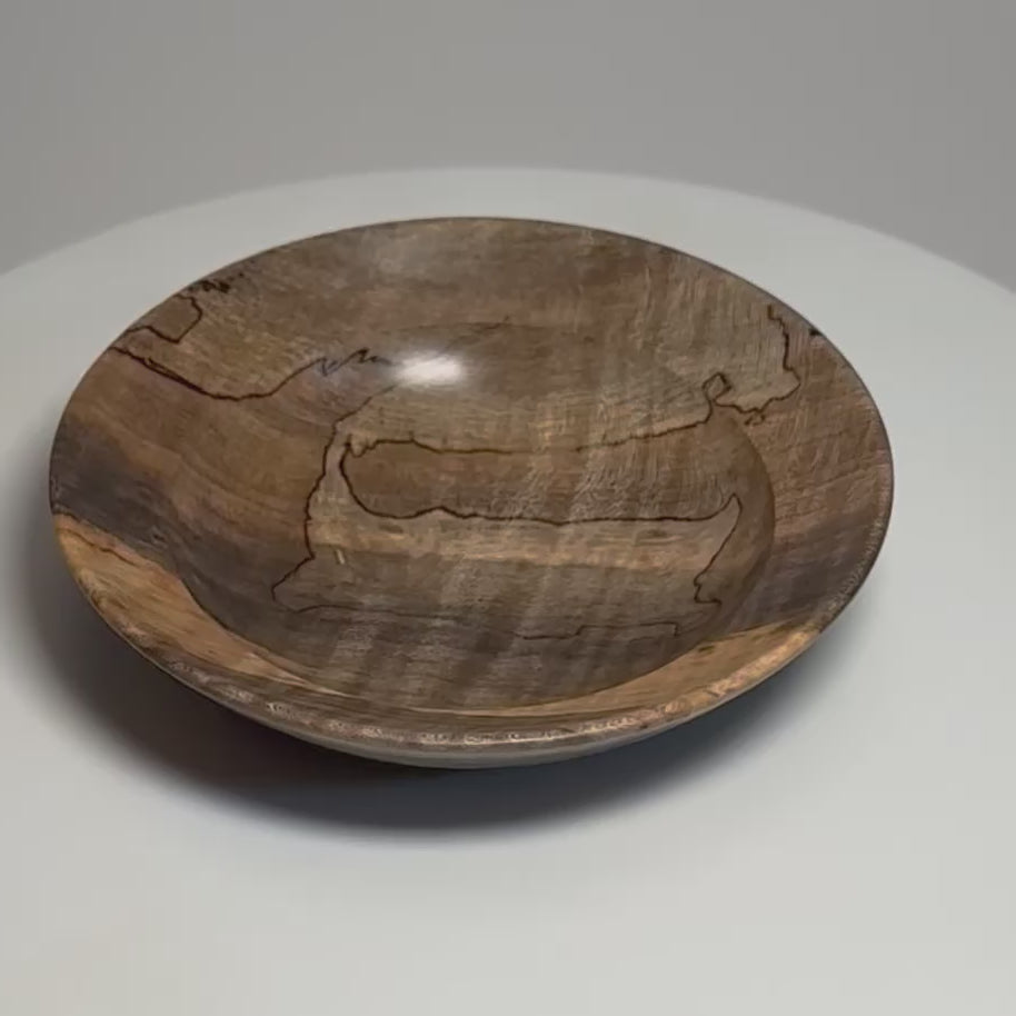 Video of a handcrafted spalted tiger maple ring dish on a rotating display with black spalting lines, earthy gold and gray-brown colors, a rounded rim and raised foot.
