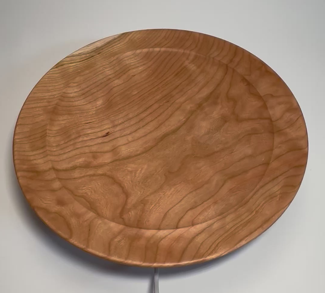 Hand-turned curly wild black cherry wooden platter rotating on display stand, showcasing warm salmon-pink curly grain and flowing figure from all angles

